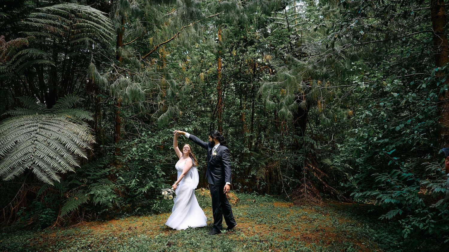 Bride and groom dancing together in lush green forest surrounded by tall ferns on wedding day.