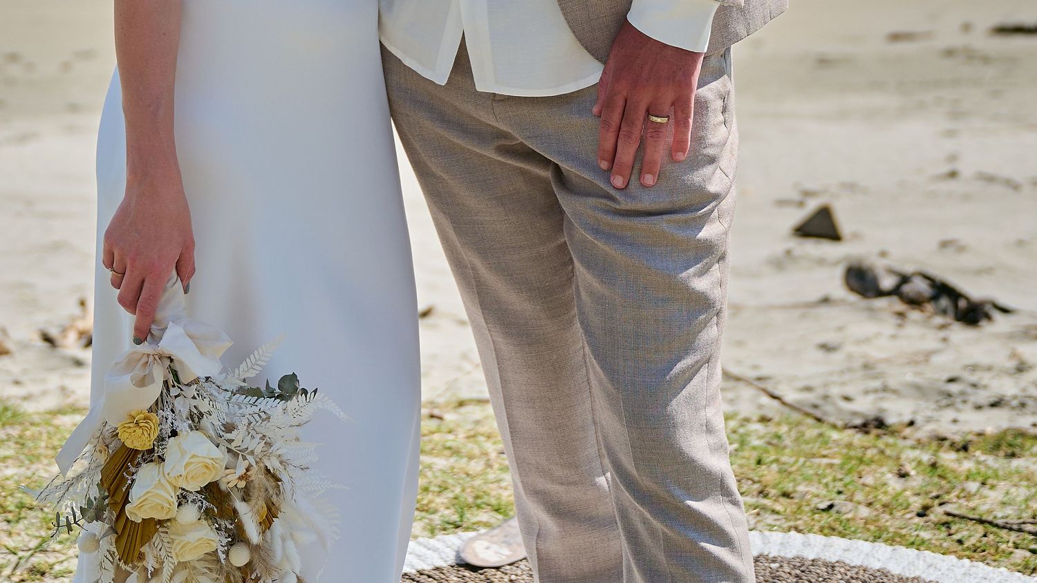 Close-up of bride and groom holding hands with a wedding bouquet of white flowers on a sandy beach.