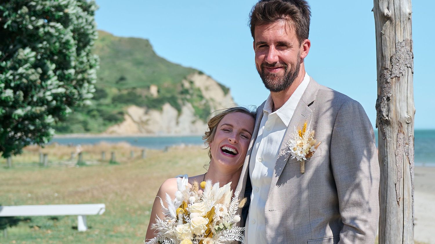 Smiling bride and groom pose together at outdoor beach wedding ceremony with white cliffs in background.