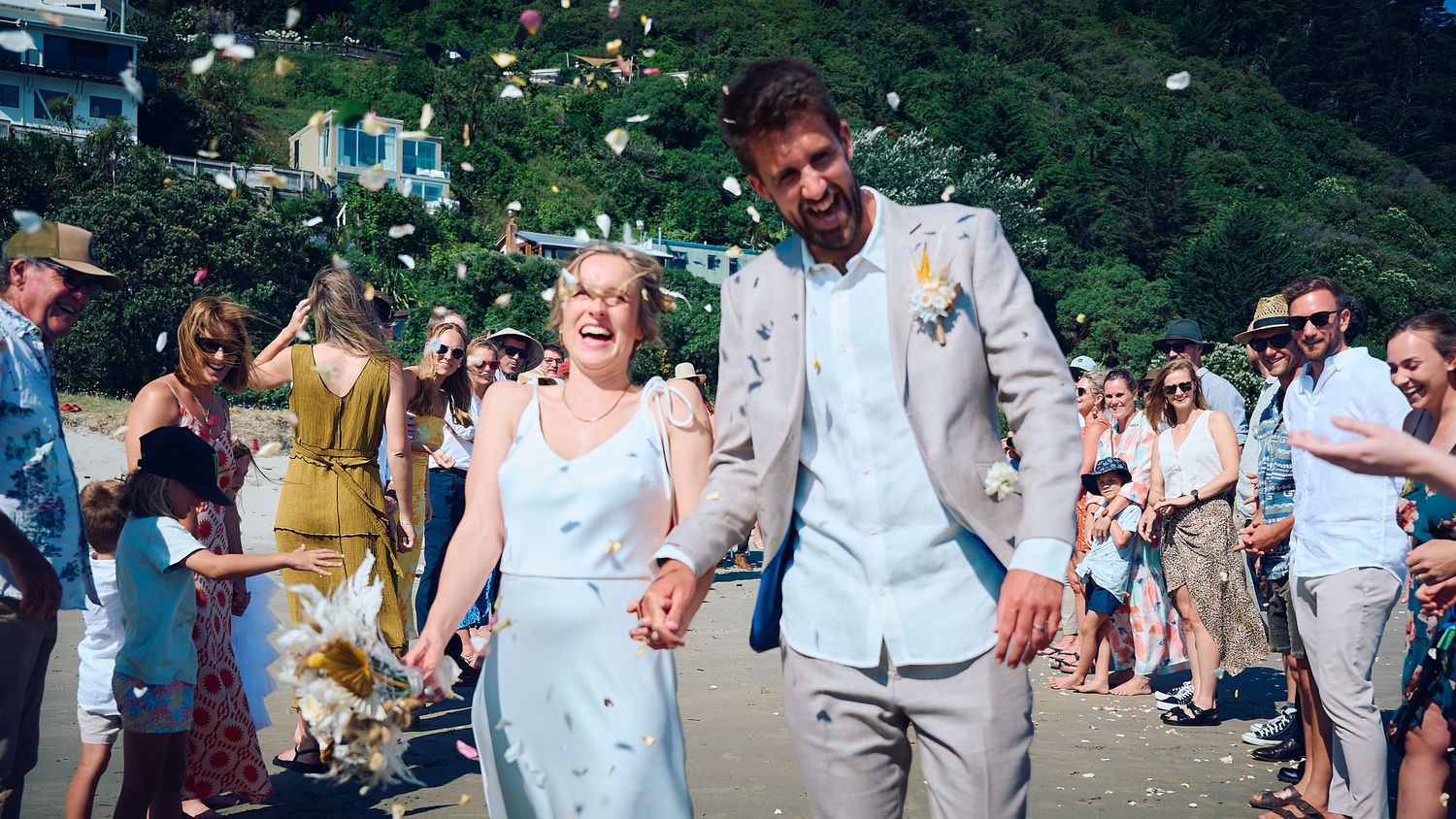Joyful newlyweds laugh as they walk through confetti shower surrounded by celebrating wedding guests on beach.