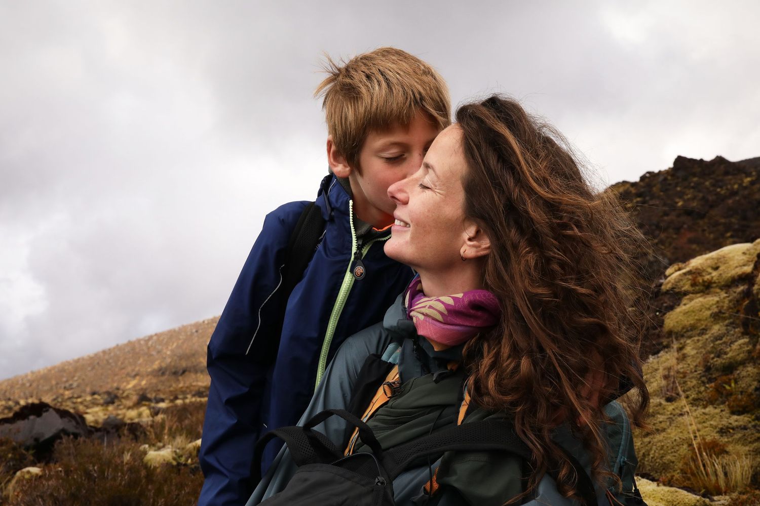 Couple sharing romantic moment on windy mountain trail with dramatic cloudy sky backdrop.