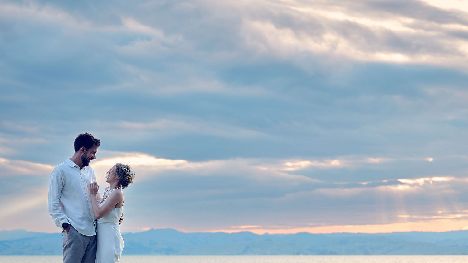 Romantic couple embraces on a beach at dusk with soft pastel sky over the ocean.