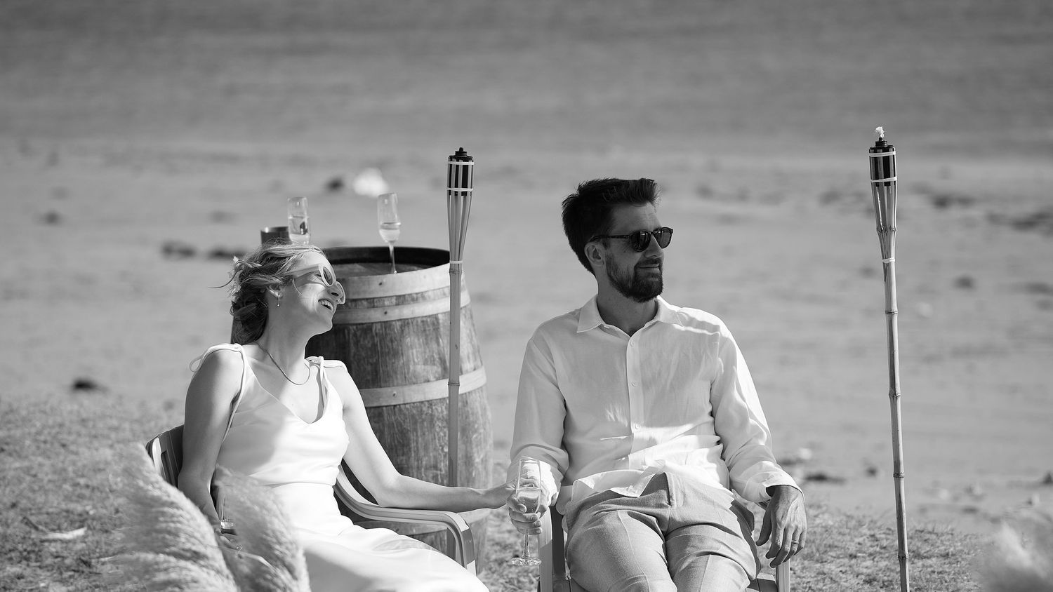 Black and white photo of bride and groom sitting relaxed beside barrel at beach wedding reception.