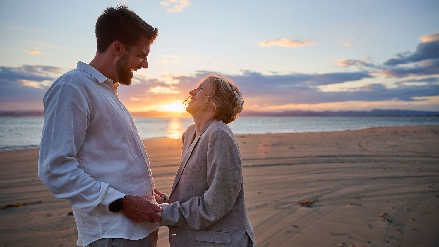 Couple holds hands and gazes at each other on a beach during a colorful sunset.