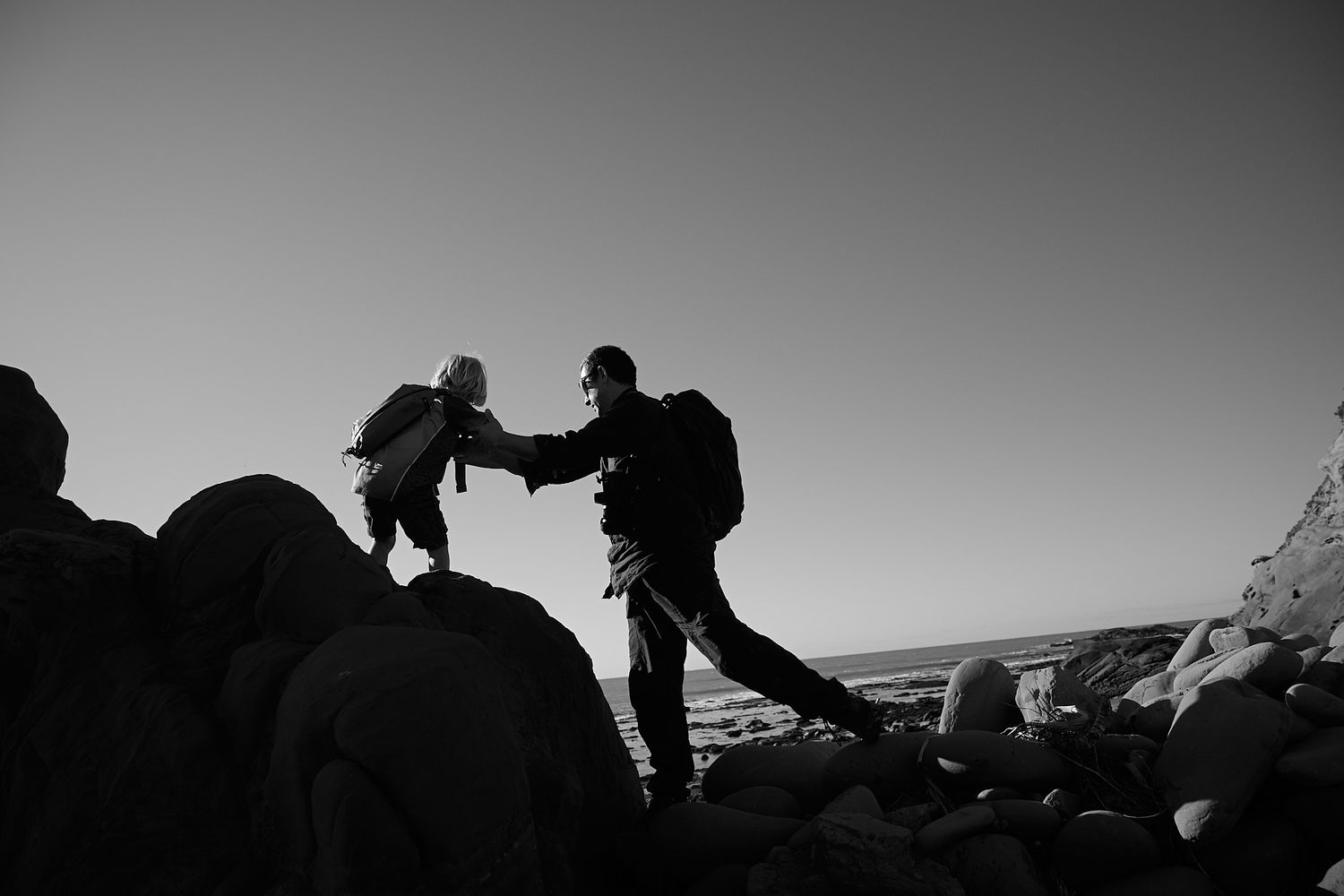 Black and white silhouette of two people climbing rocks near the ocean at dusk.