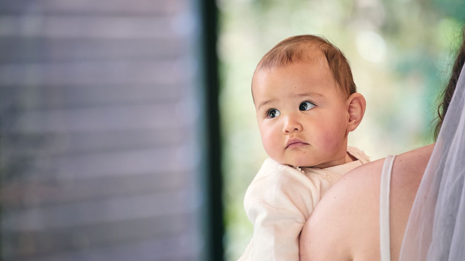 Adorable Asian baby looking over mother's shoulder in soft natural light.