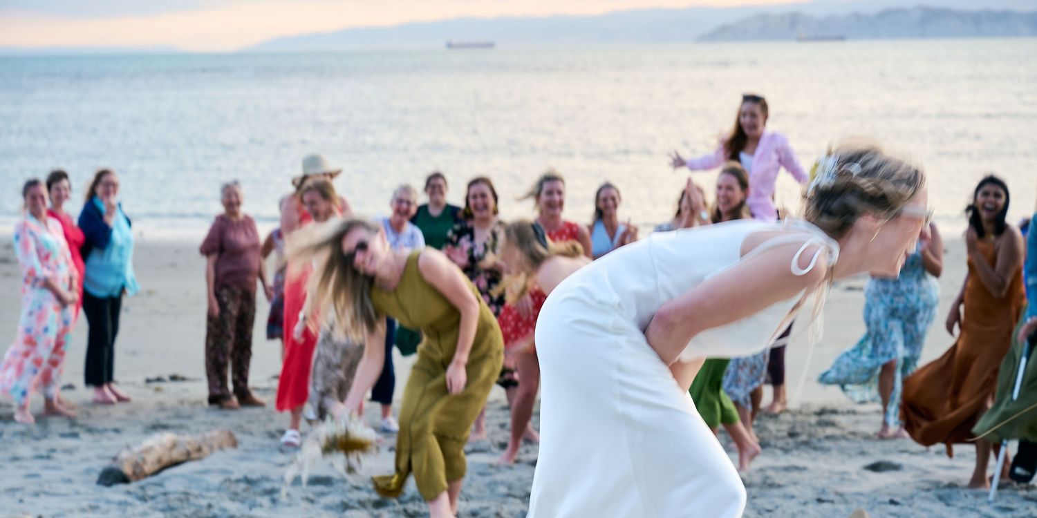 Bride bends forward tossing bouquet to excited guests at beach wedding.