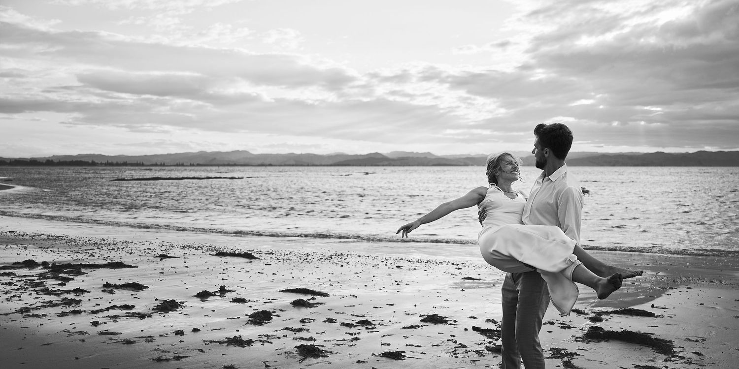 Black and white photo of a man carrying a woman on a beach with ocean backdrop.