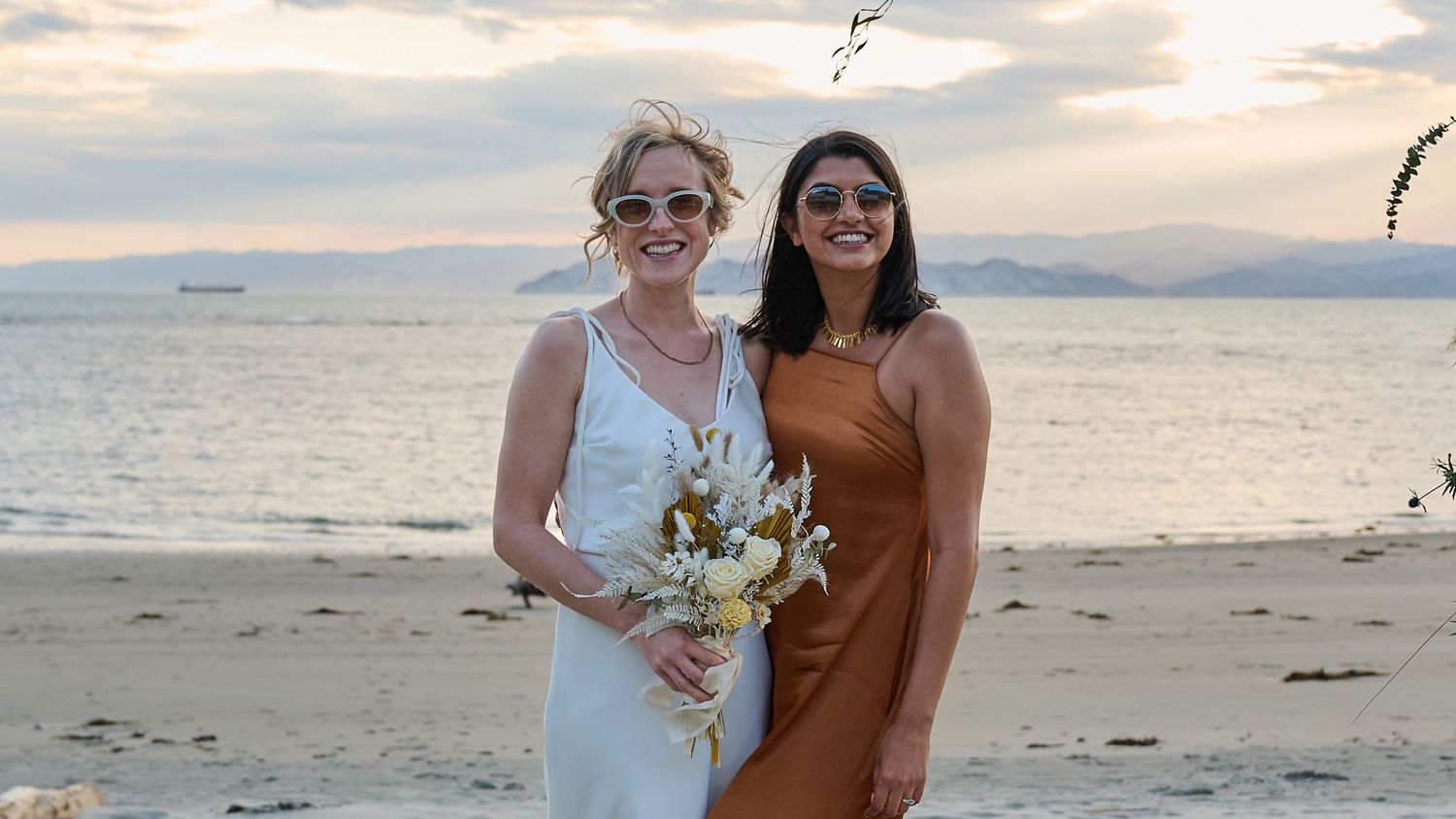 Two women smiling together on a beach at sunset, one in a white wedding dress holding a bouquet.