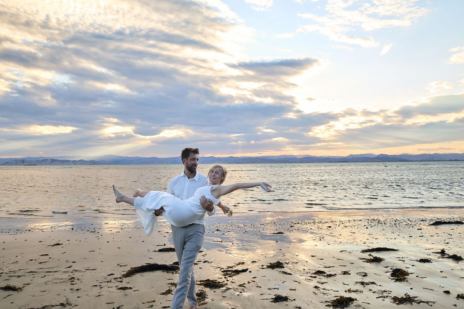 Man carries woman in his arms on a beach at sunset with dramatic cloudy sky.