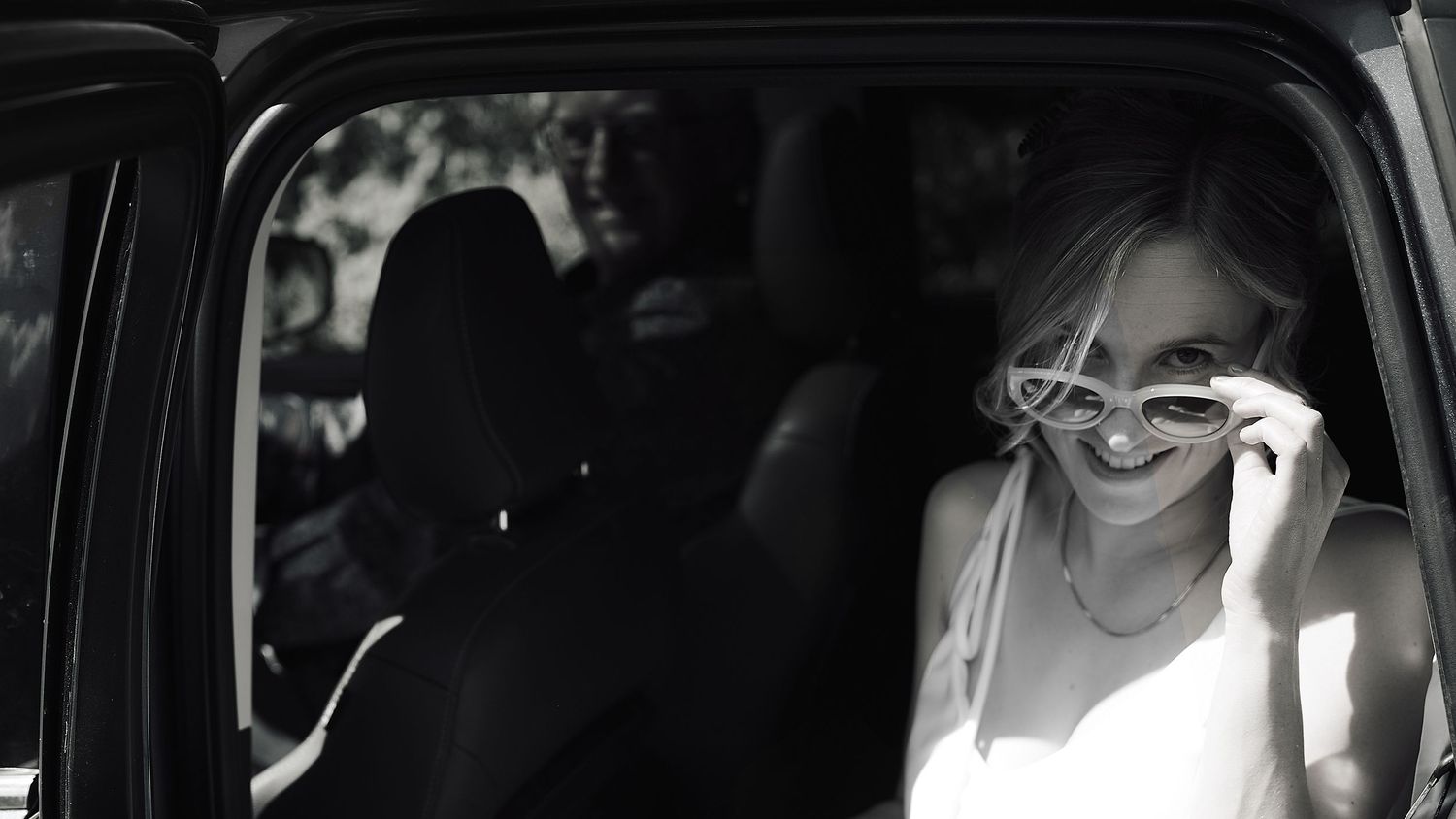 Smiling bride in white dress adjusts sunglasses while sitting in a car.