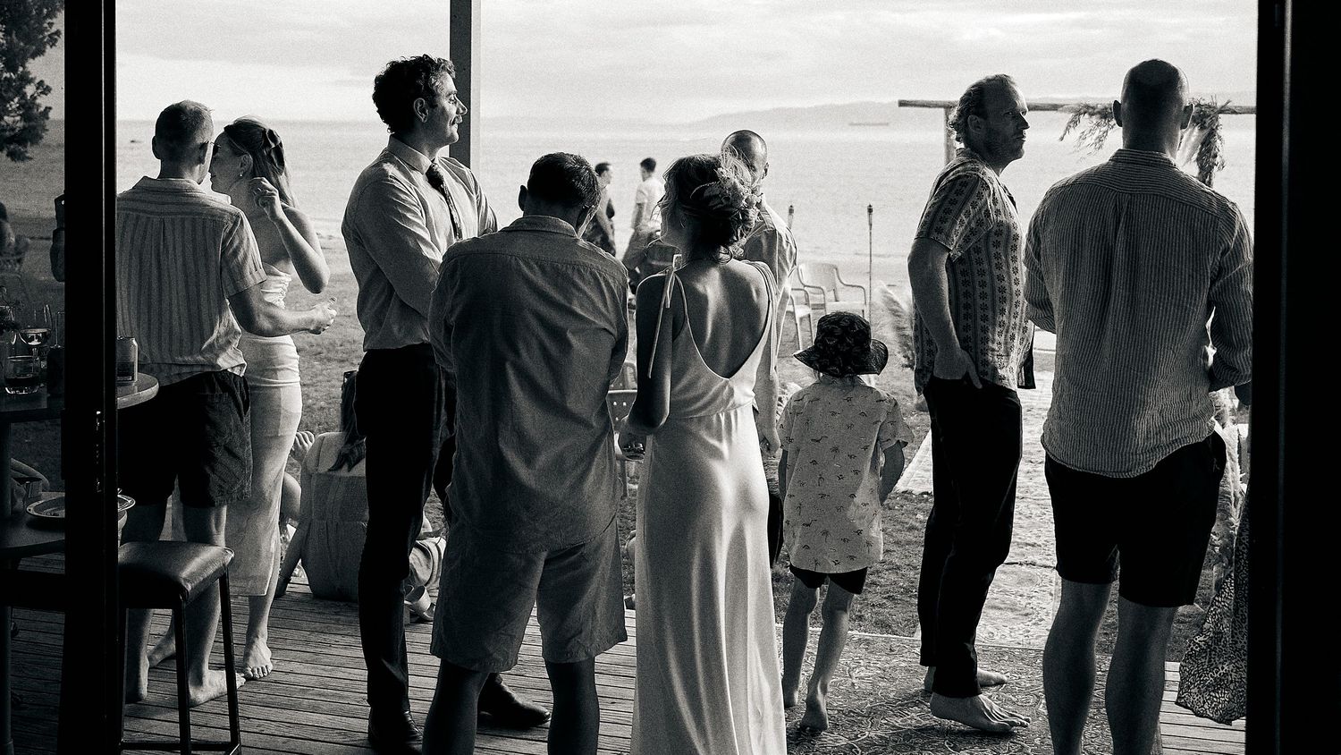 Black and white photo of wedding guests silhouetted against large windows with an ocean view.