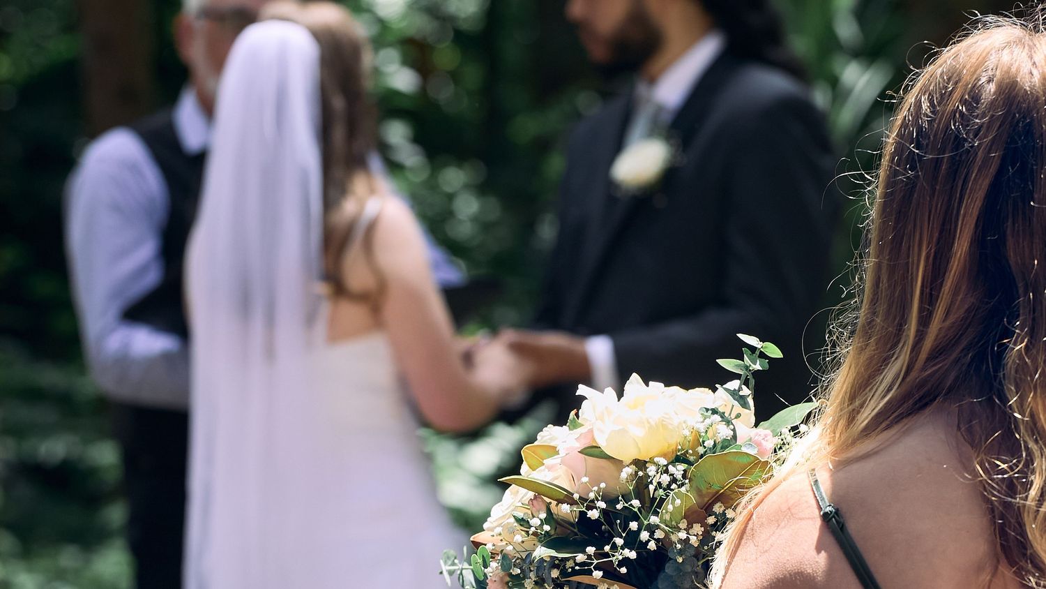 Wedding guest holding bouquet of white flowers watching outdoor ceremony.