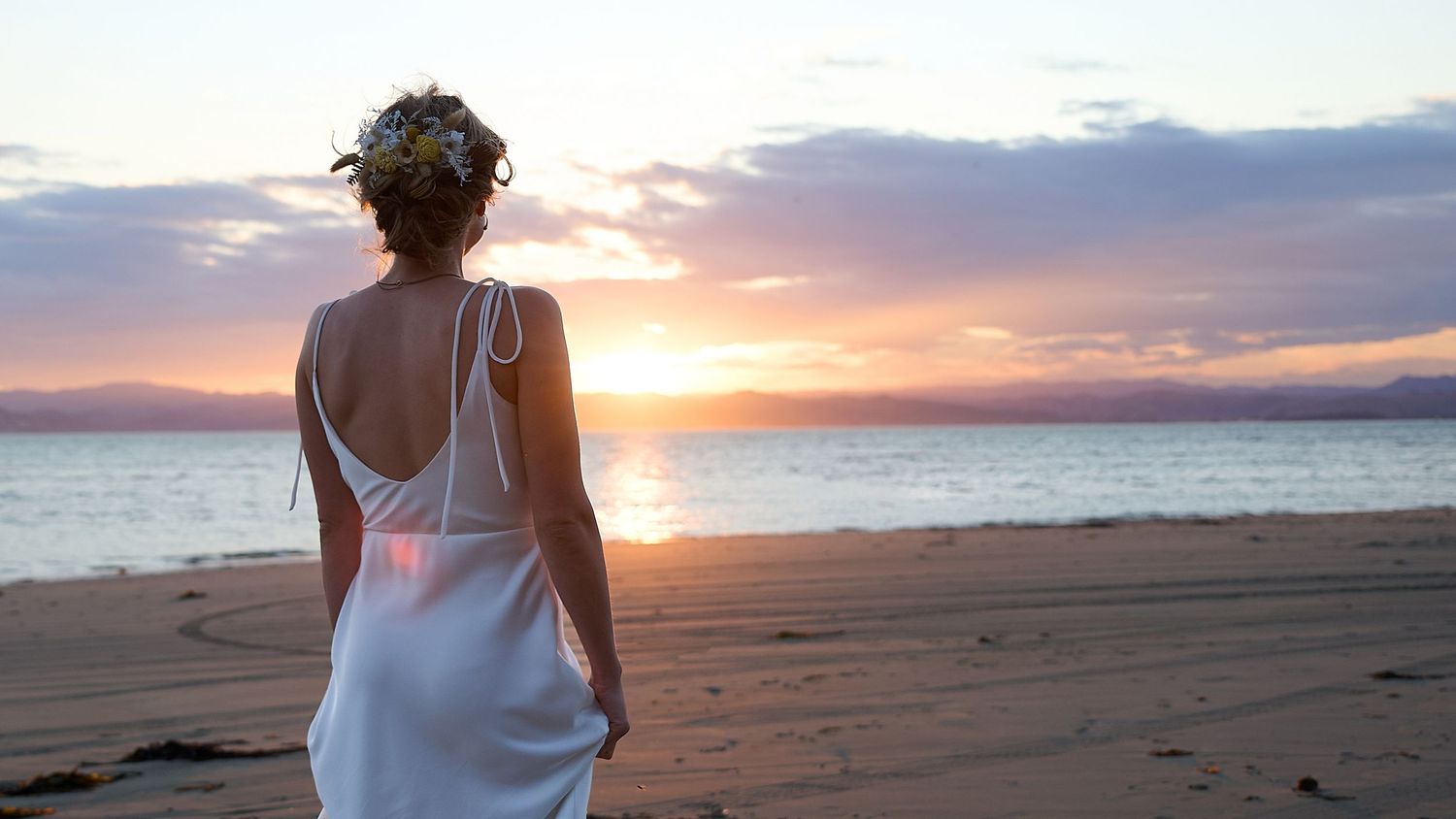 Bride in a backless white gown standing alone on a beach watching a colorful sunset over the ocean.