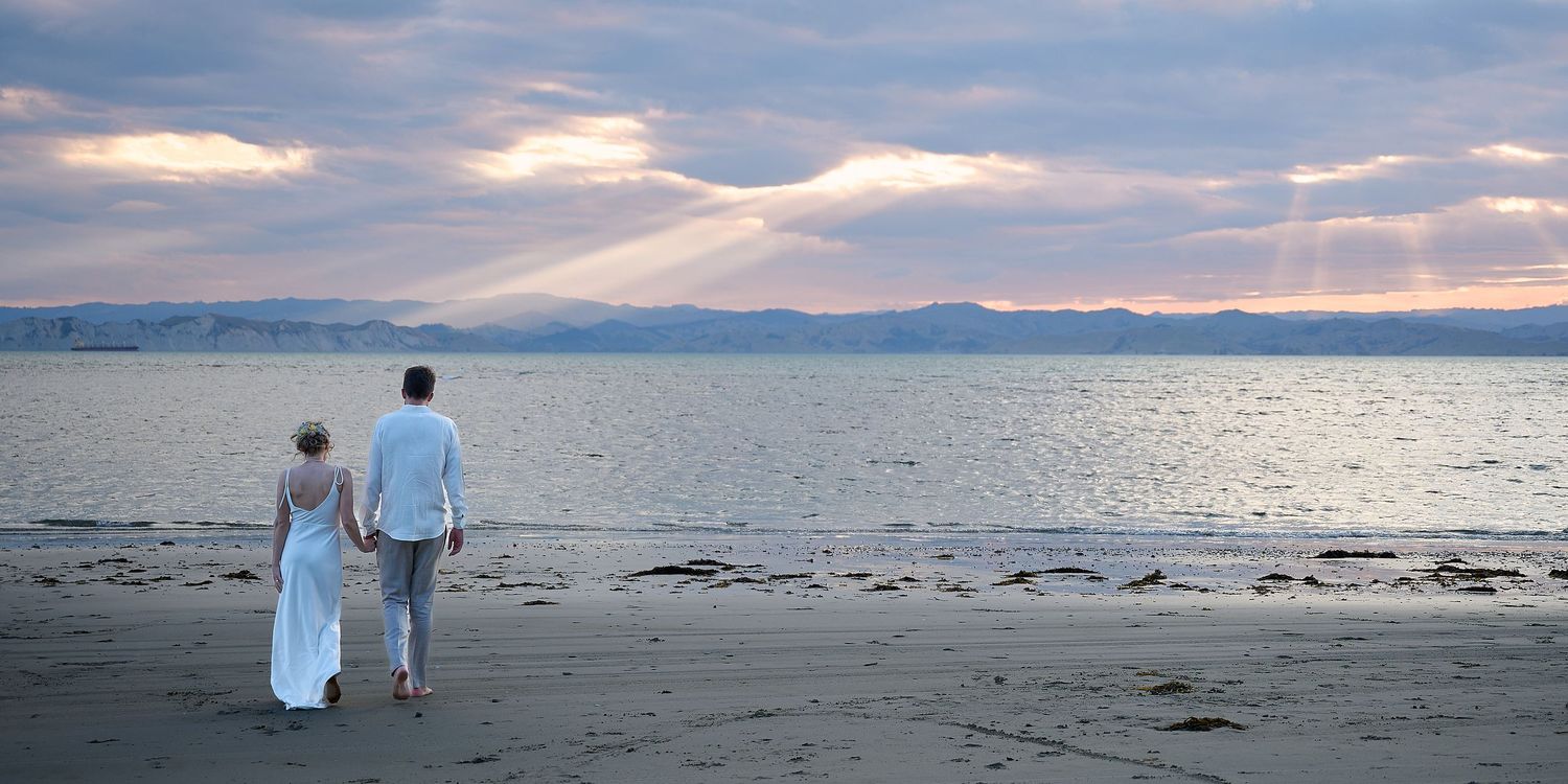 Couple in wedding attire walking hand in hand on a serene beach at sunset with dramatic sky.