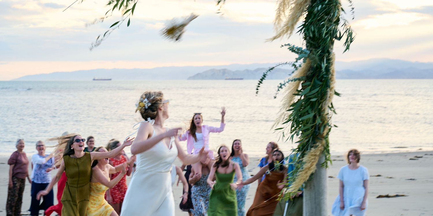 Bride tosses bouquet to guests at sunset beach wedding ceremony under floral arch.