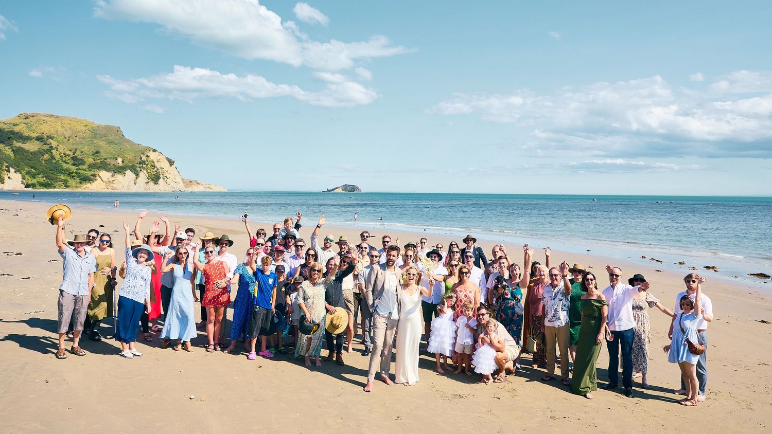 Large wedding party group photo on a sunny beach with a rocky island visible in the background.