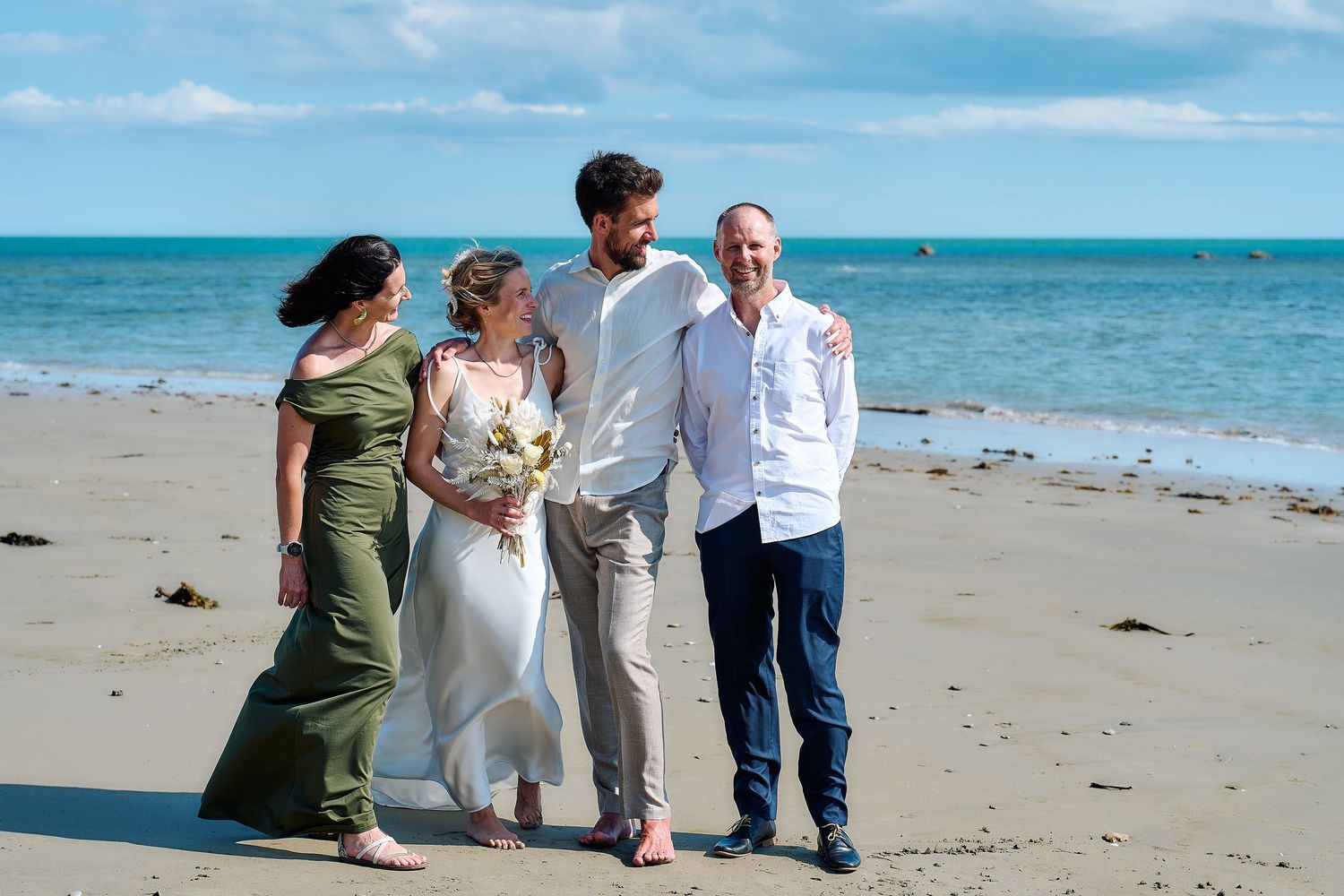 Newlywed couple poses with two family members for portrait on sunny sandy beach after wedding.