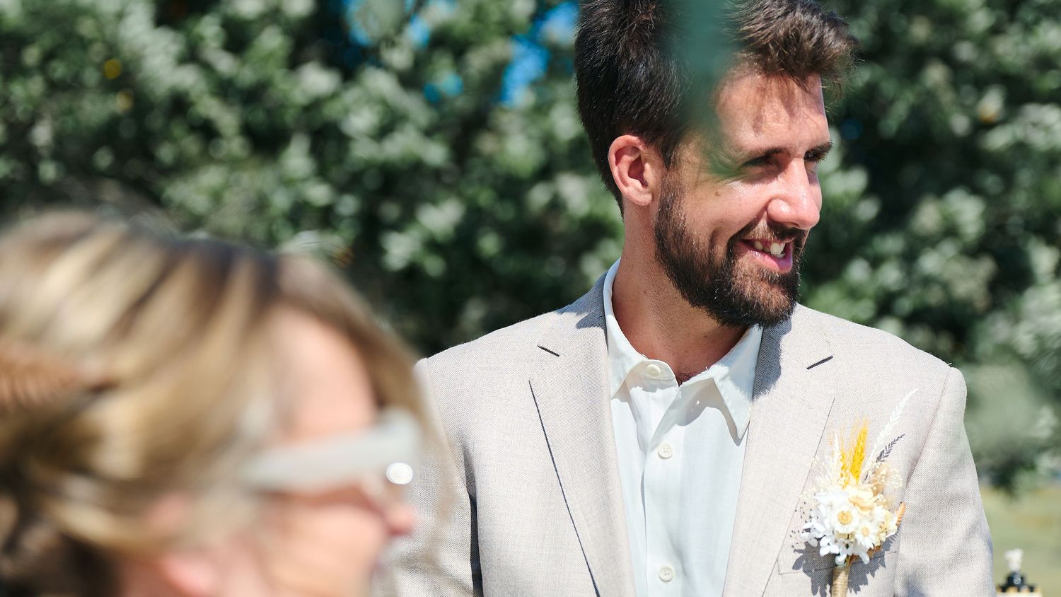 Smiling groom in a beige suit with a floral boutonniere standing outdoors during a wedding ceremony.