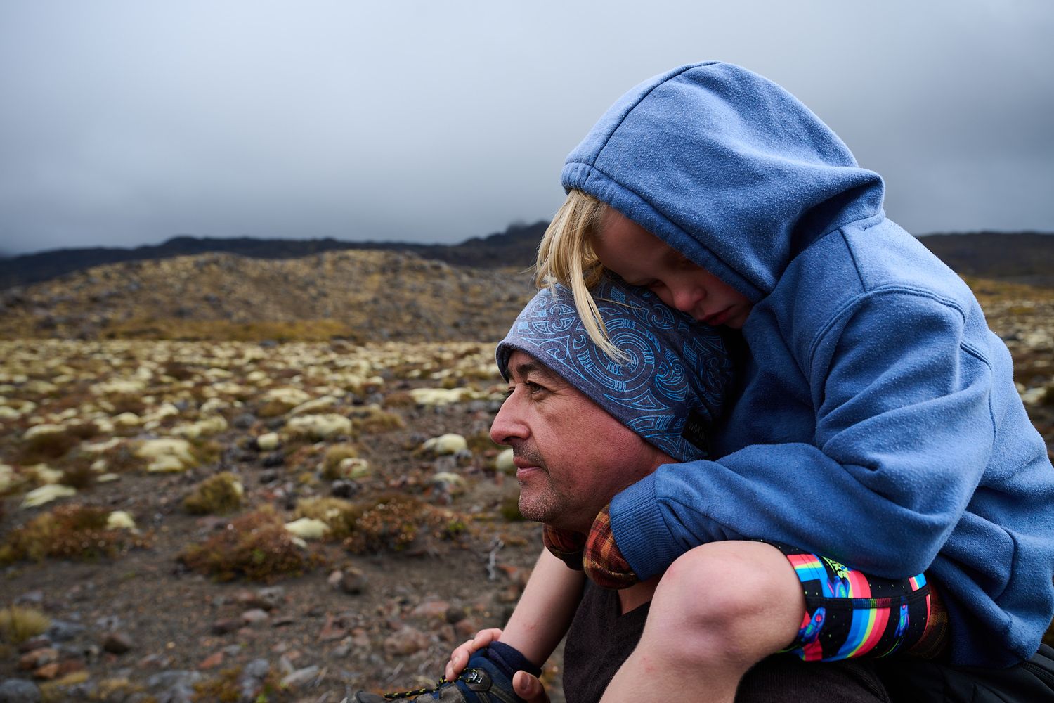 Adult carrying a child on their back while exploring a rugged volcanic landscape on a cloudy day.