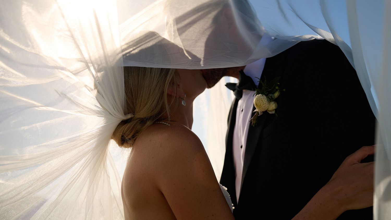 Couple kissing under billowing white veil with sunlight streaming through on wedding day.
