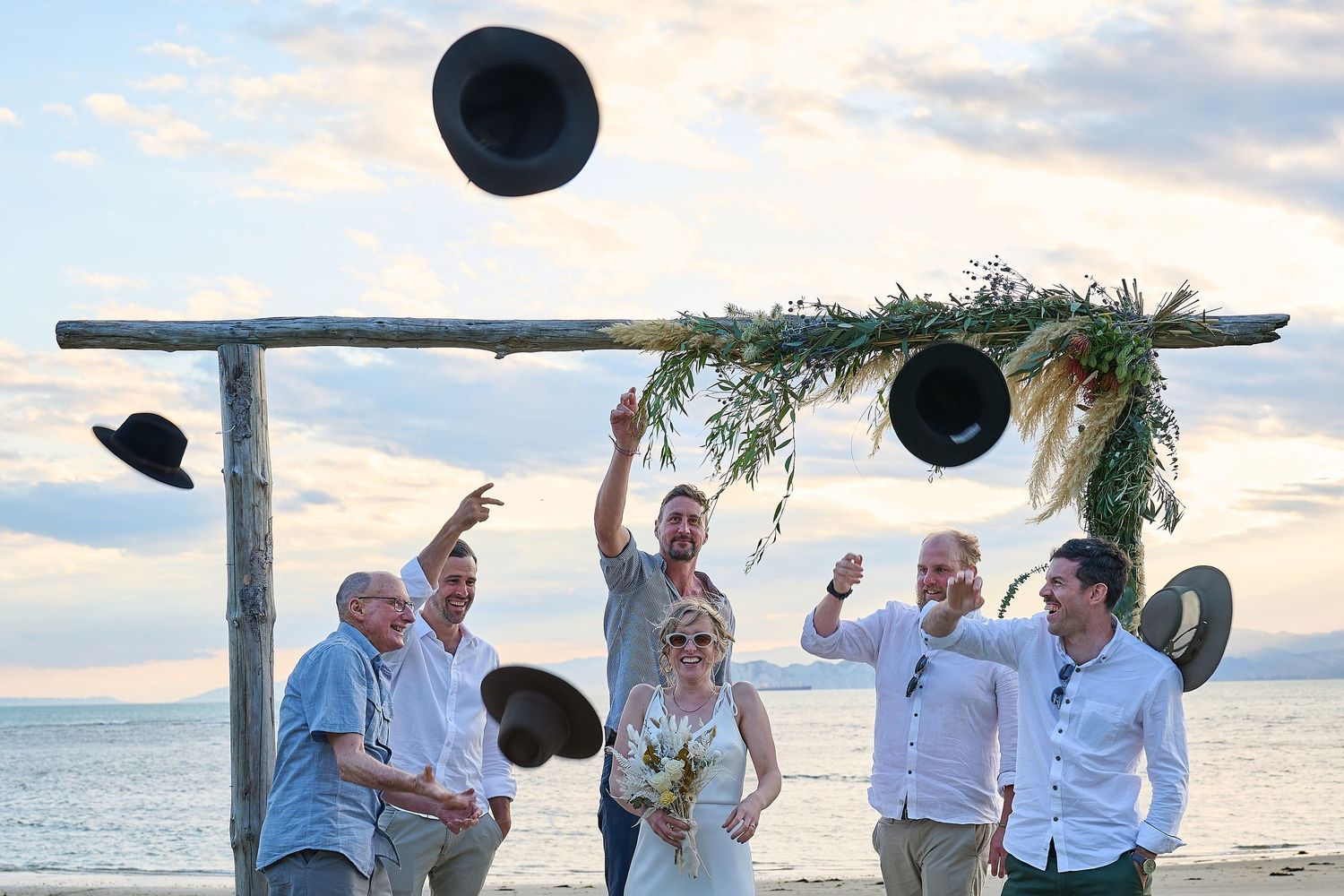 Wedding party tosses hats in the air celebrating at sunset beach ceremony.