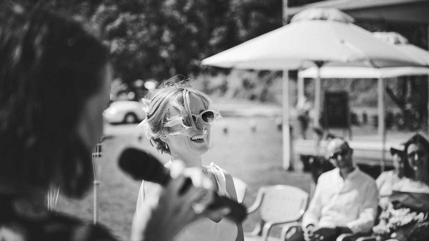 A laughing bride in sunglasses enjoys a speech at an outdoor wedding reception in black and white.
