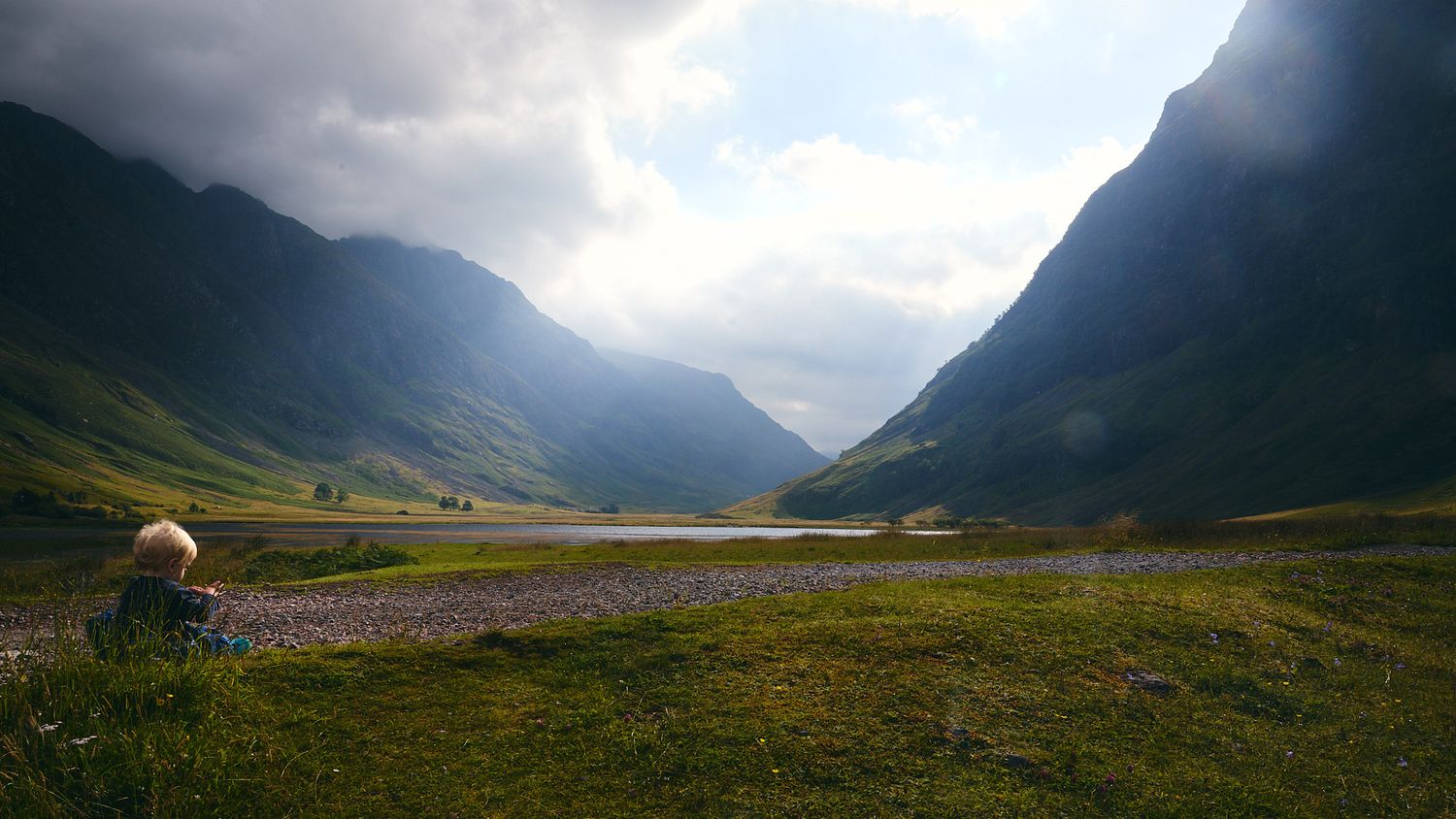 Person sitting alone gazing at a stunning green valley with a lake between misty mountains.