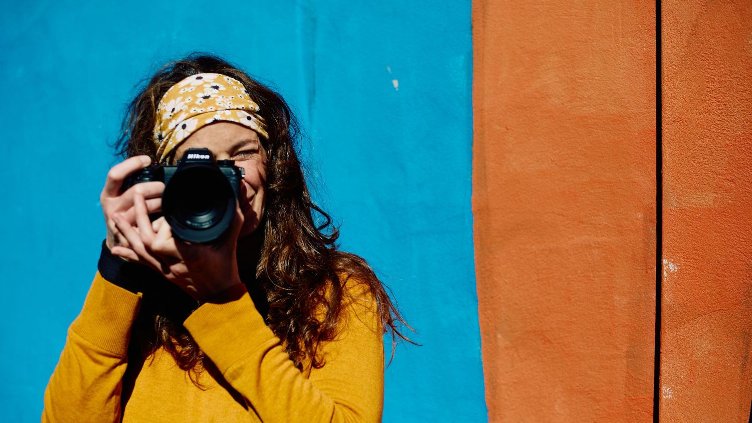 Woman in yellow sweater with bandana photographing with Nikon camera against colorful blue and orange wall.