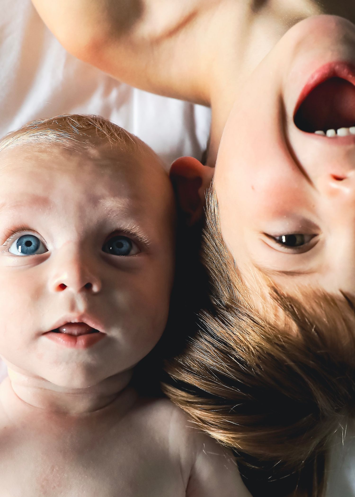 Close-up of a blue-eyed baby lying beside a laughing child on a white surface.