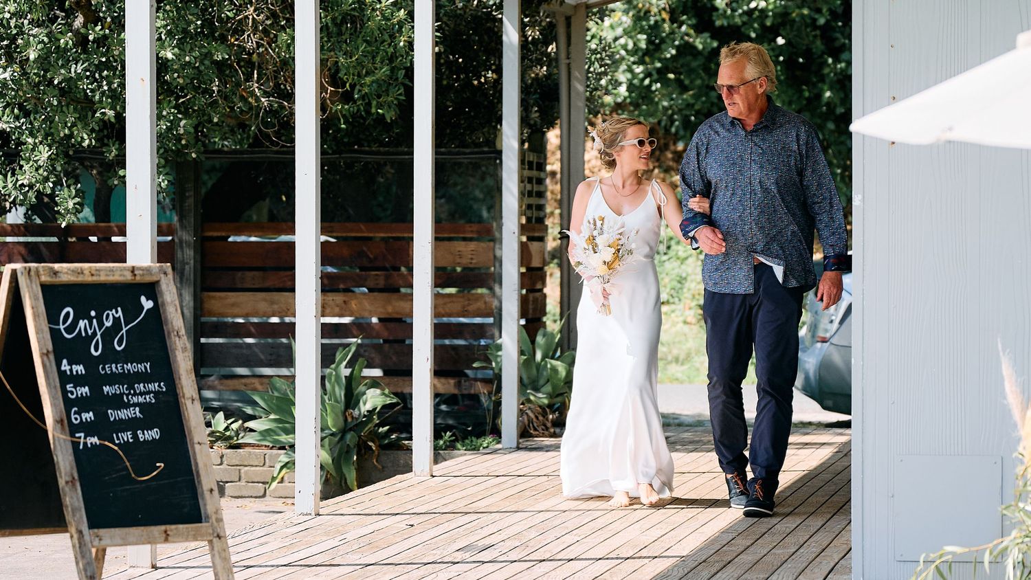 A bride in a white dress walks with an older man along a wooden deck past a chalkboard sign.