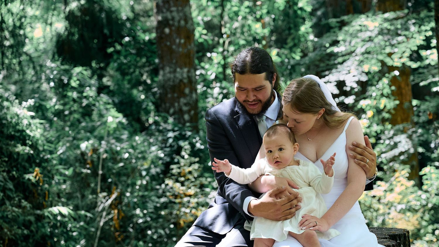 Smiling bride and groom crouching down with their baby in a lush green forest setting.