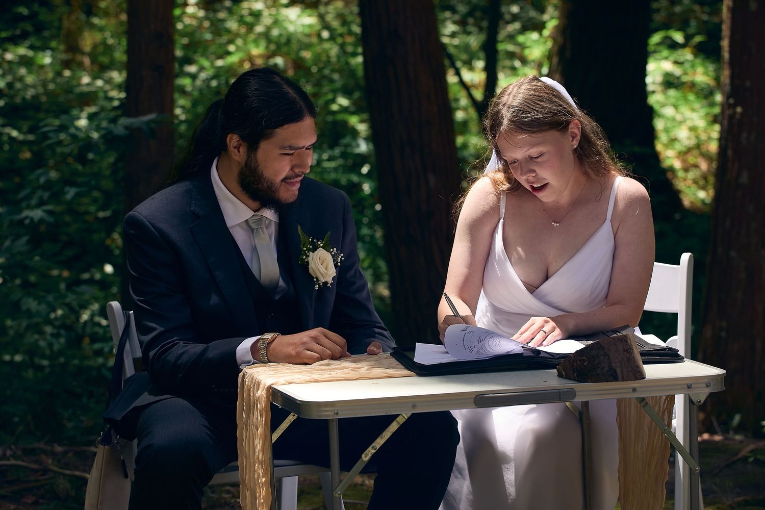 Bride and groom laughing together while signing wedding documents at outdoor table in forest setting.