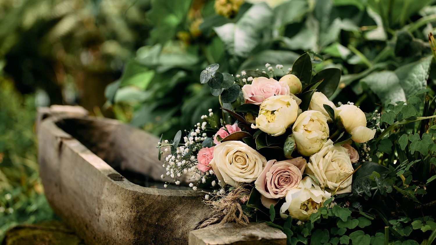 Elegant bridal bouquet of cream and pink roses resting in a rustic wooden bowl outdoors.
