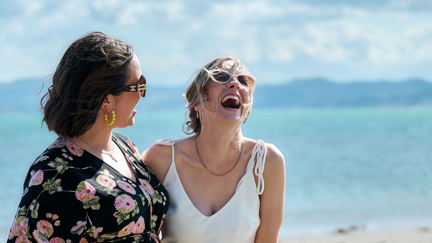 Two women laughing joyfully on a beach, one in a white dress, on a sunny day with blue ocean waters behind them.
