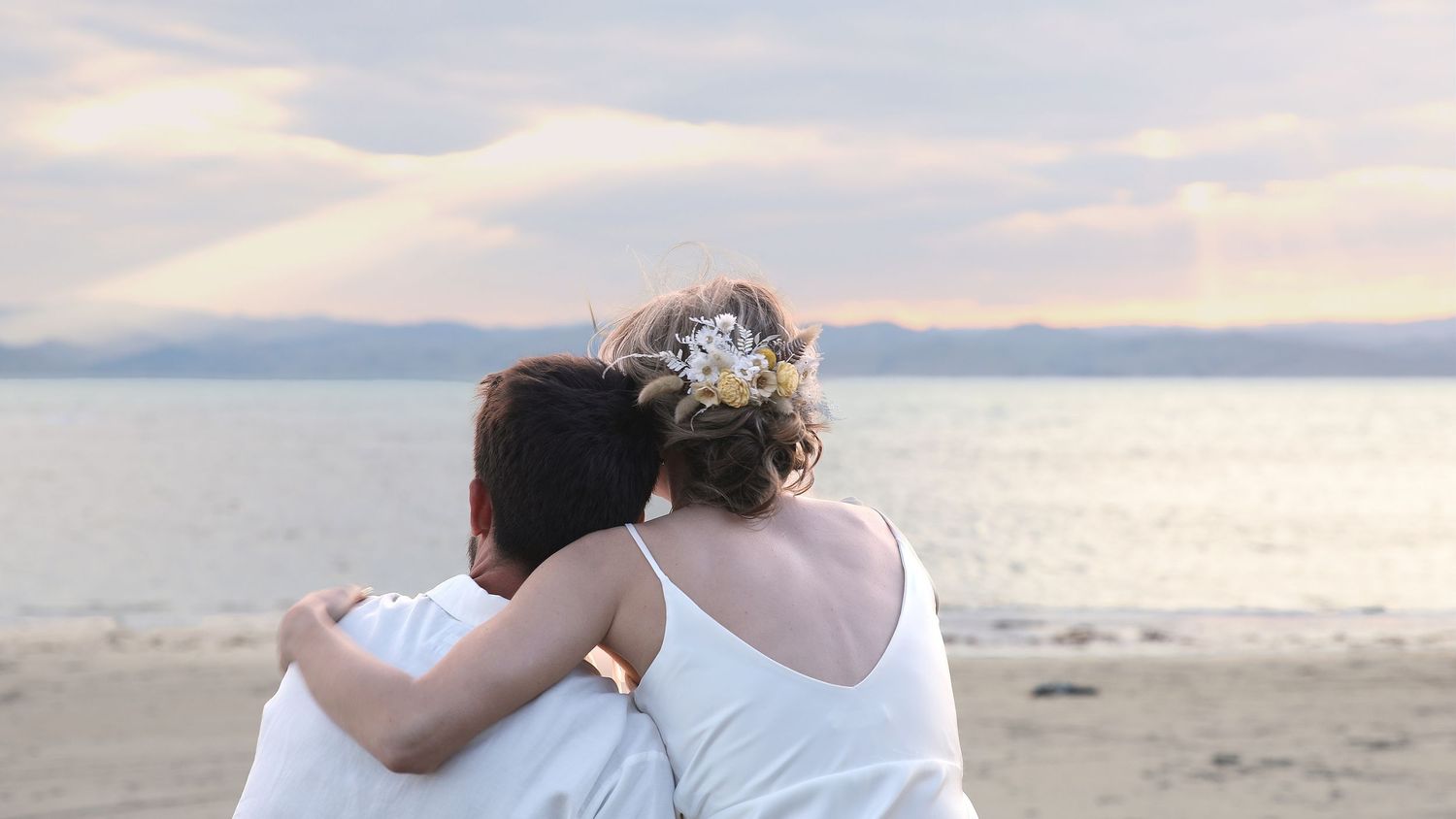 Bride and groom embrace facing sunset ocean at romantic beach wedding.