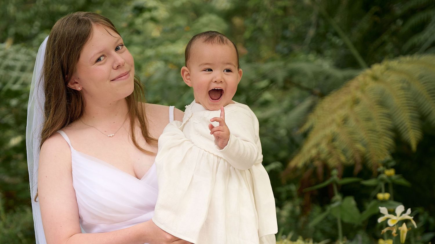 Smiling bride holding laughing baby girl in cream dress at outdoor wedding.