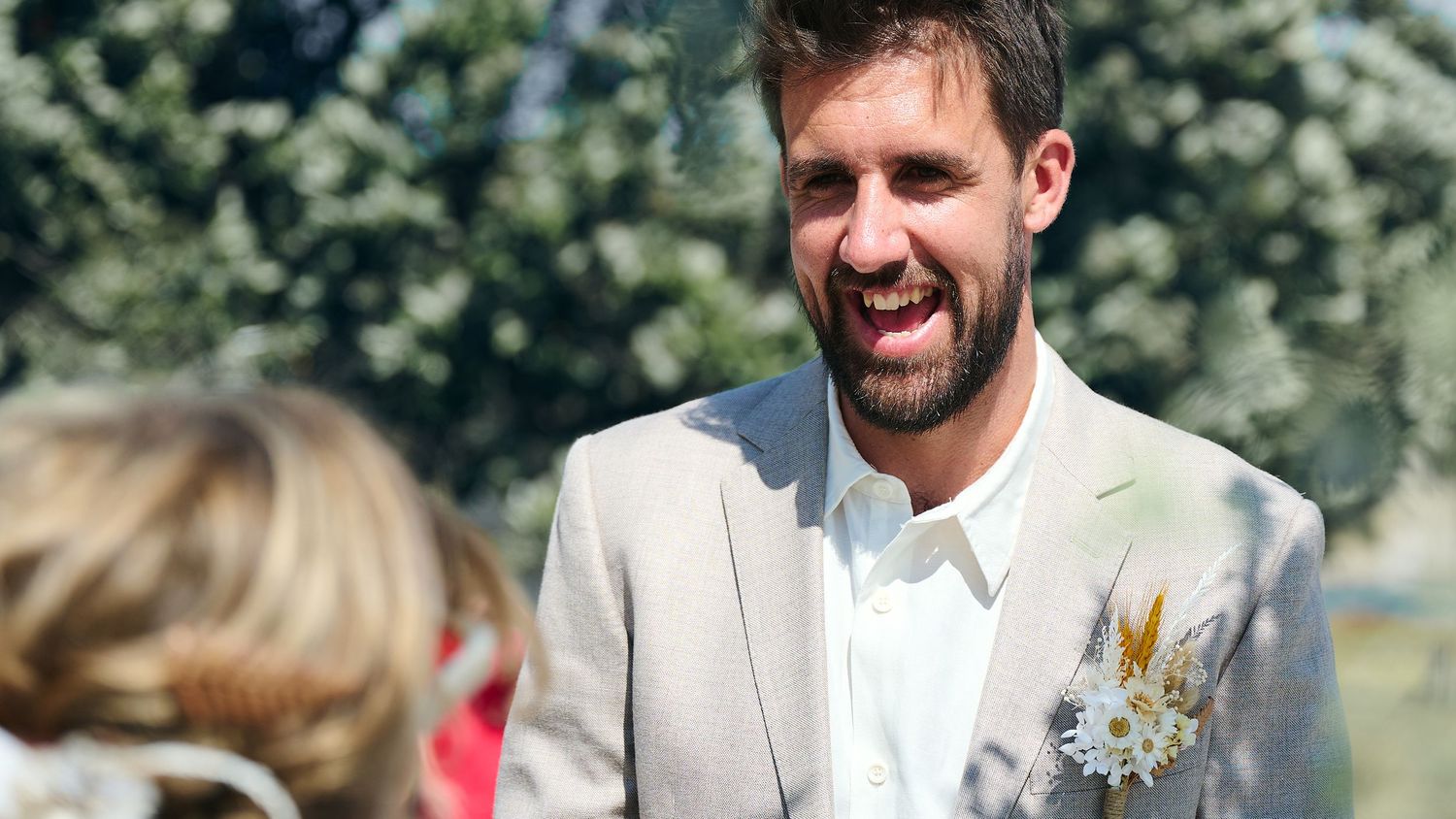 Happy groom in a light grey suit with boutonniere laughing during an outdoor wedding ceremony on a sunny day.