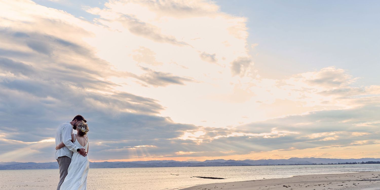 Bride and groom share romantic embrace on beach under dramatic cloudy sky.