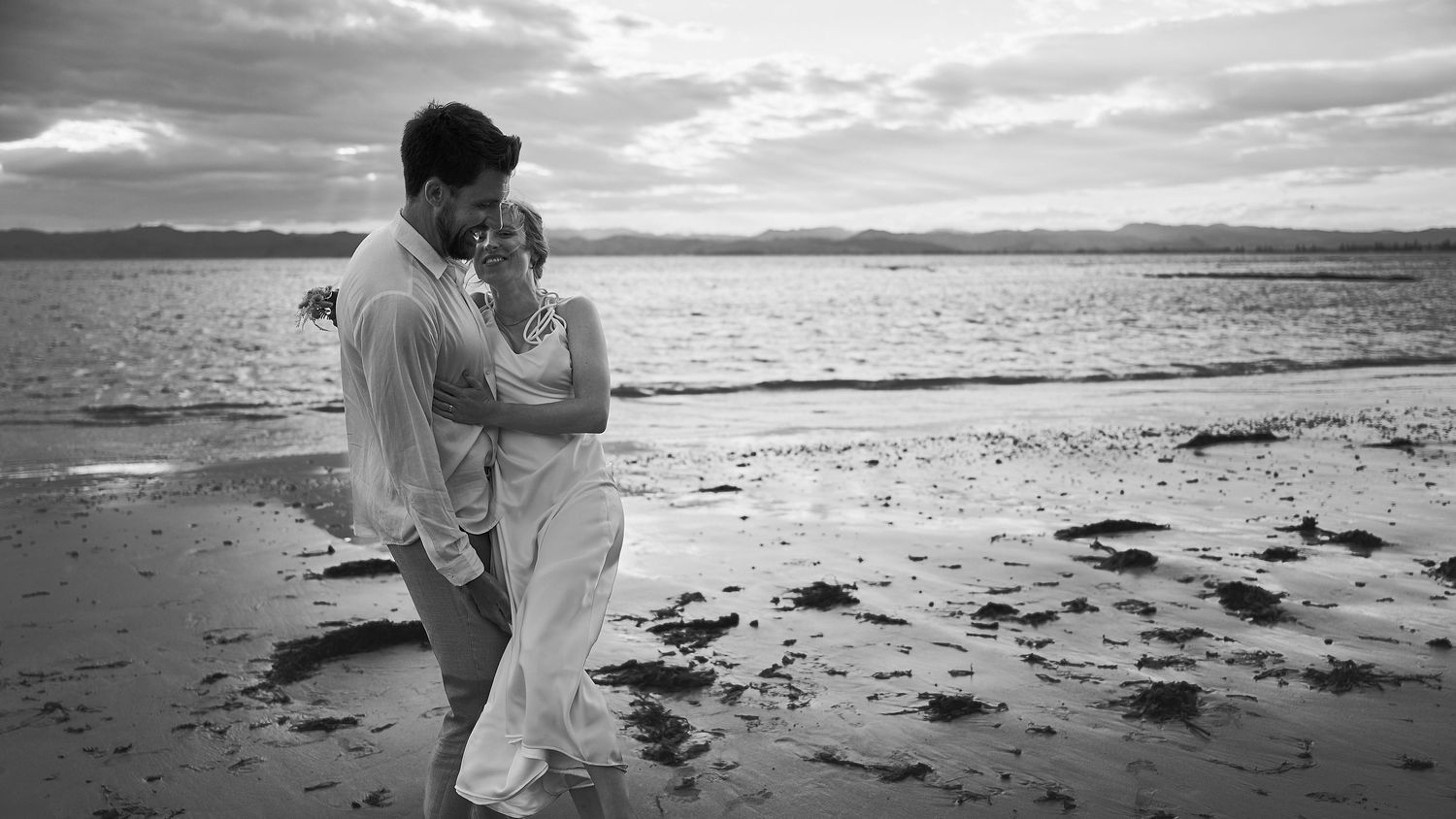Black and white image of a couple embracing on a sandy beach with ocean waves nearby.