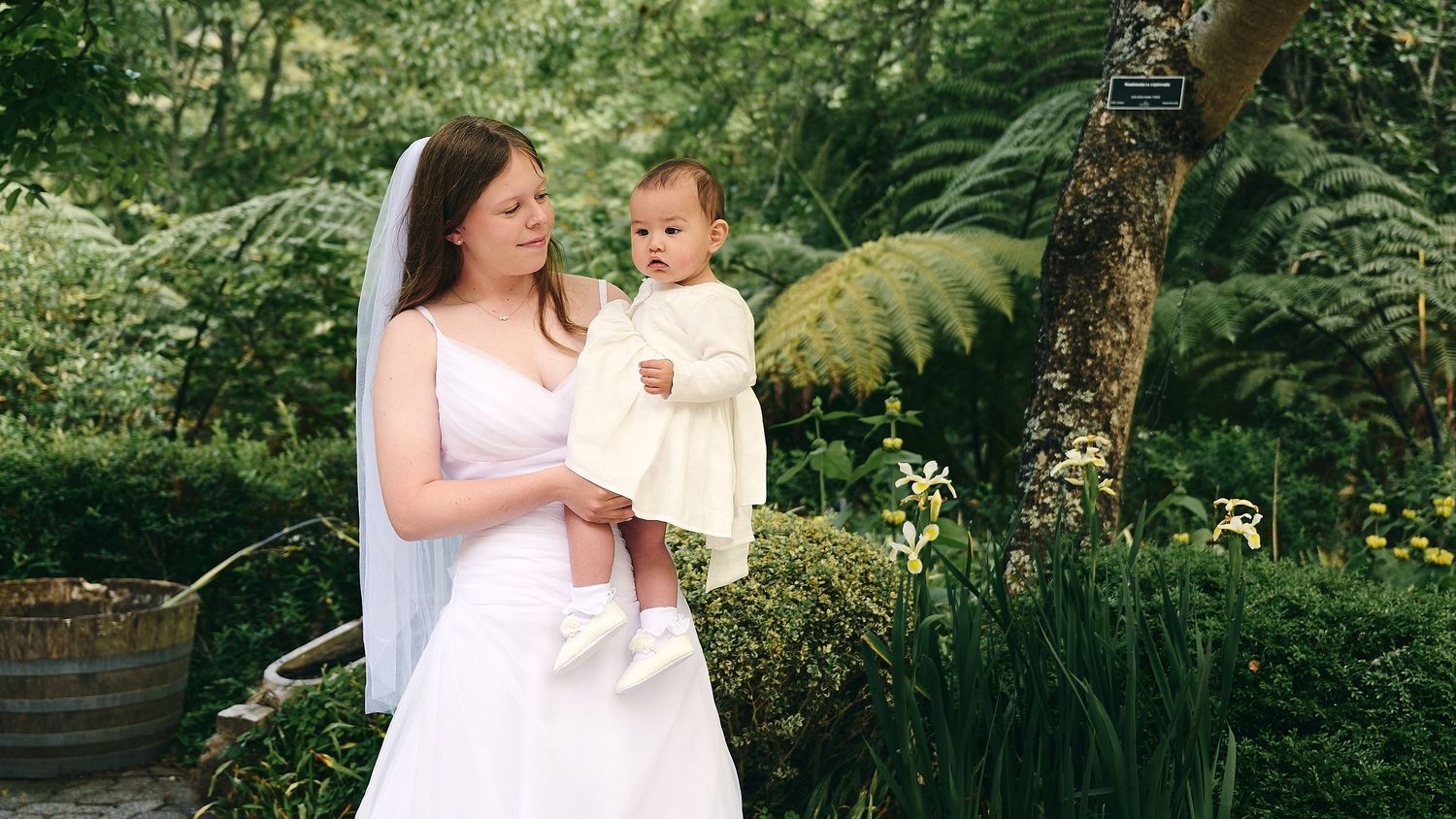 Bride in white wedding dress holding baby girl in lush garden setting.