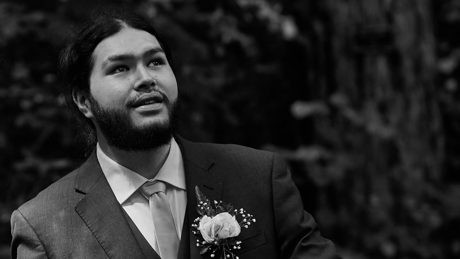 Black and white portrait of a bearded groom in a suit with a boutonniere looking upward.