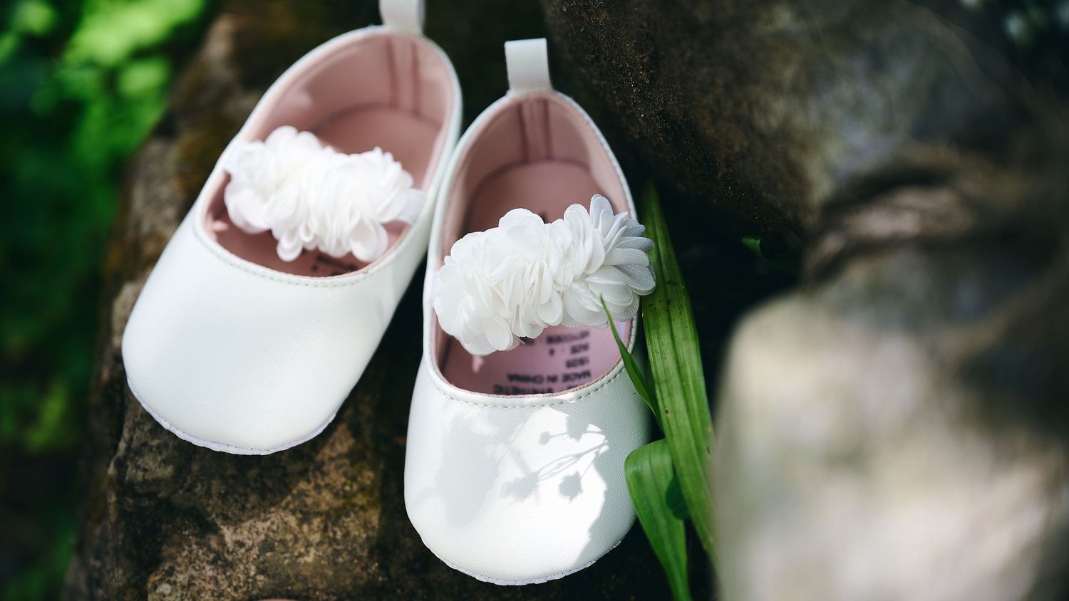 White baby girl shoes with floral detail resting on a tree bark surface.