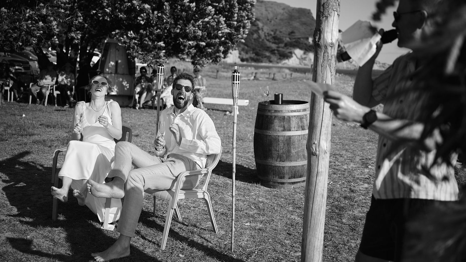 Black and white photo of wedding couple seated outdoors while guest plays music beside wooden barrel.