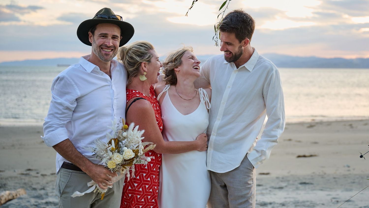 A joyful group of four celebrating a beach wedding at sunset, with the bride holding a white bouquet.
