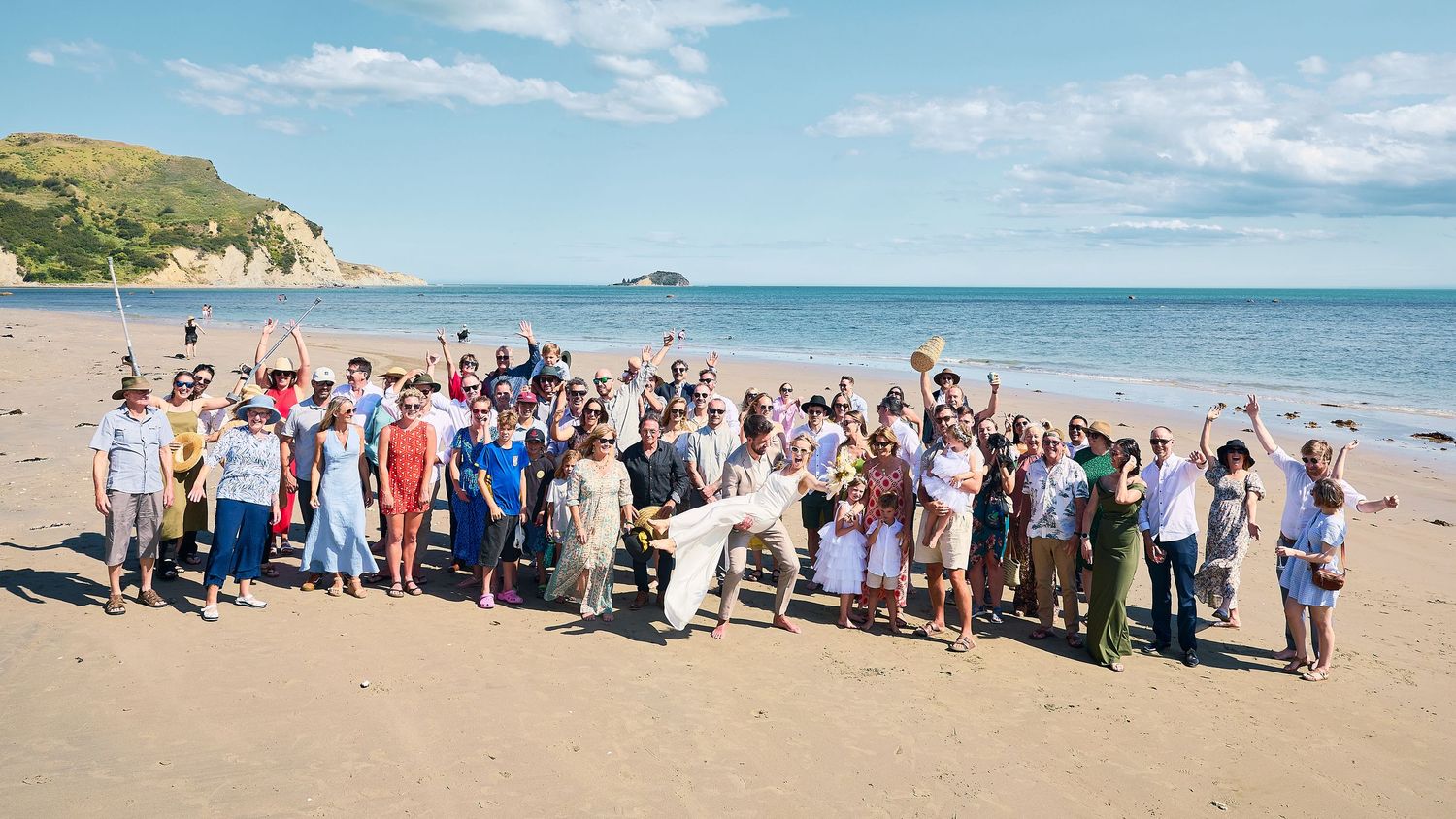 Cheerful wedding guests posing together on a sandy beach with blue skies and a rocky island backdrop.