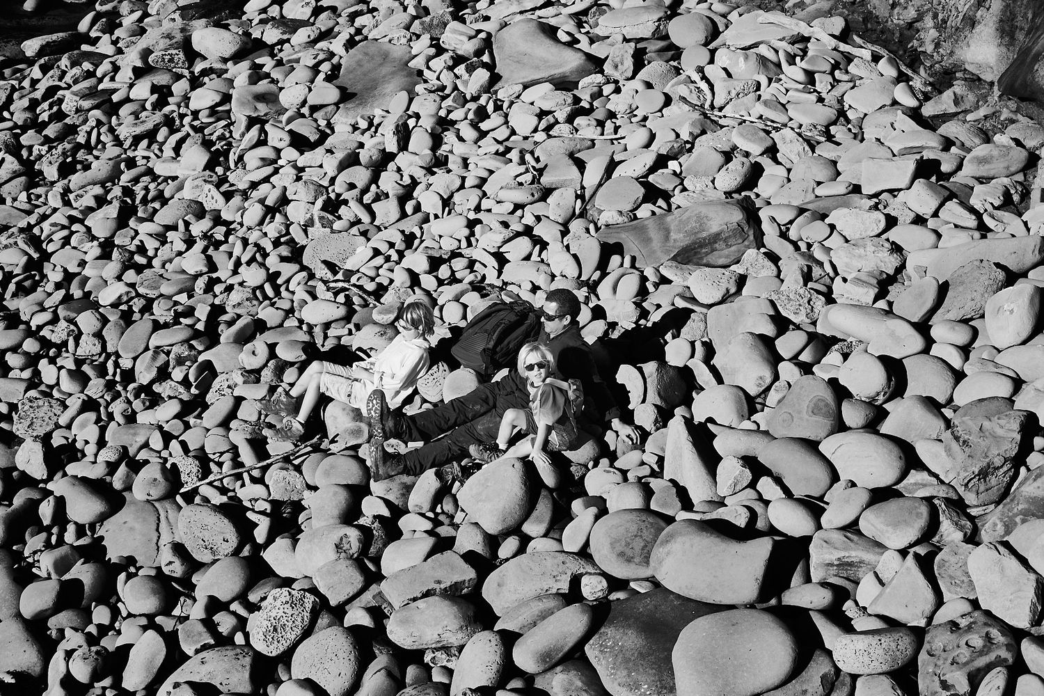 Aerial black and white photo of two people lying among large rounded rocks on a rocky beach.