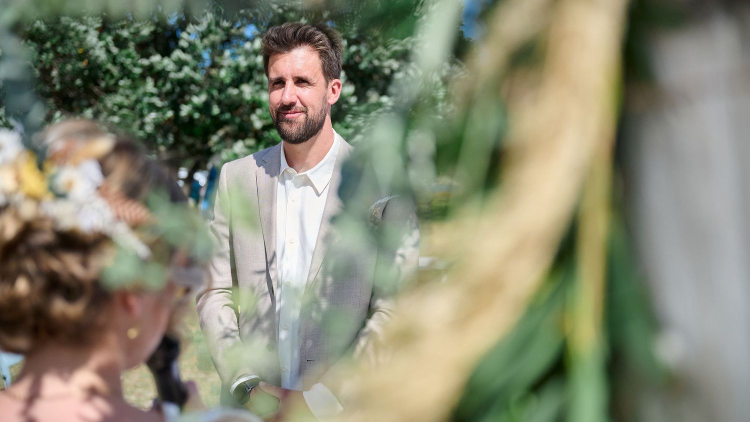 Groom in a grey suit standing attentively during an outdoor wedding ceremony surrounded by lush green foliage.
