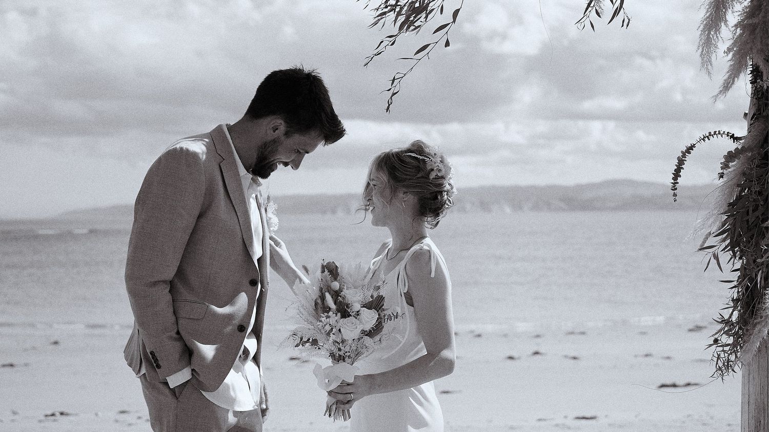 Black and white romantic photo of bride and groom facing each other on beach during wedding ceremony.