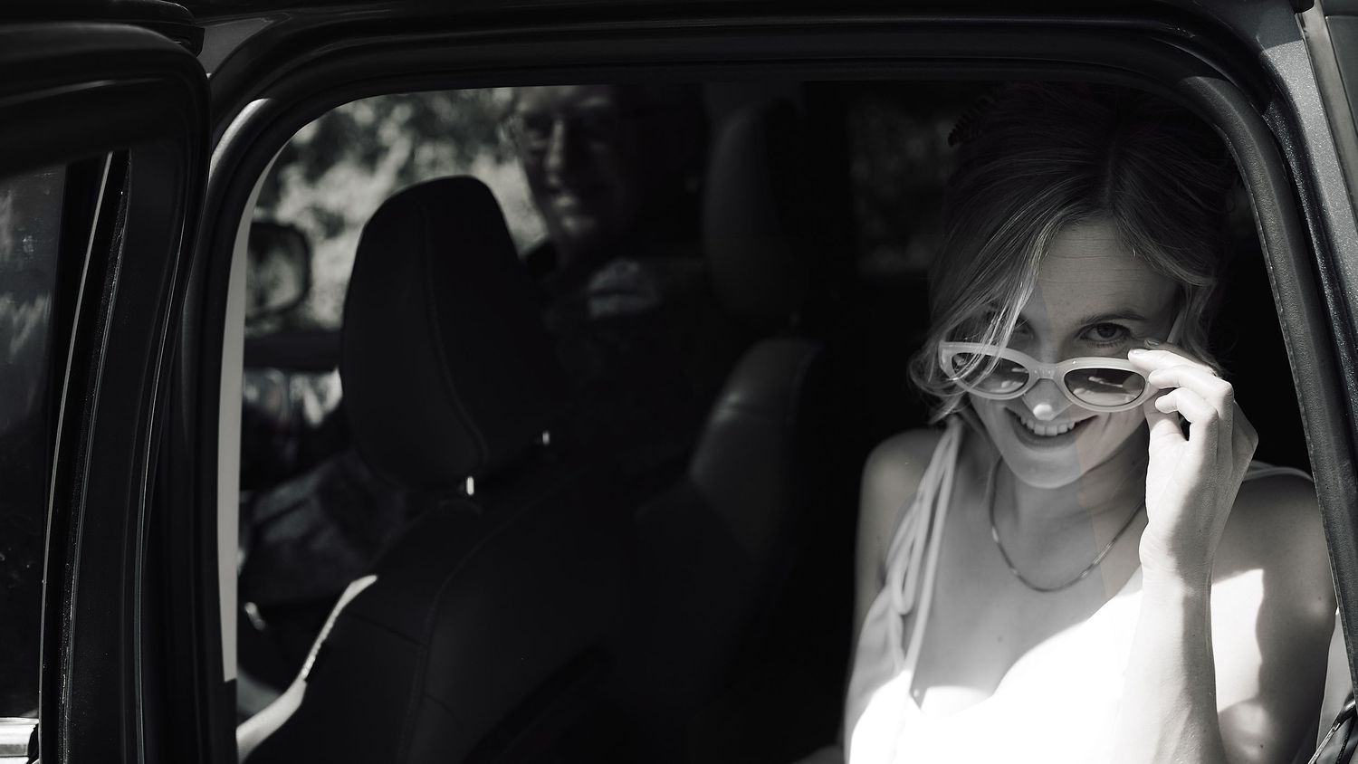 A smiling bride in white adjusting her sunglasses inside a car in a classic black and white photo.